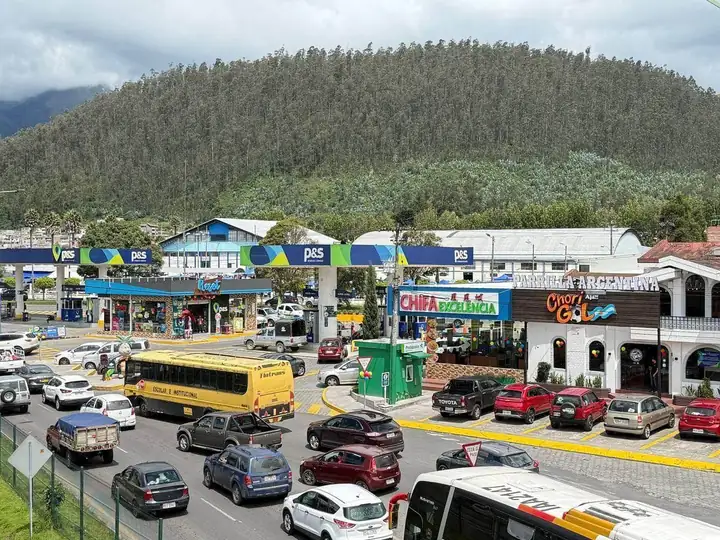 Vista de Plaza Redondel desde la Panamericana Norte: gasolinera, locales de comida y estacionamiento junto a un cerro verde, en Otavalo